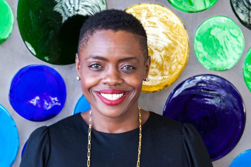 Rafat Fields smiling in a black blouse in front of a colorful glass-disc wall.
